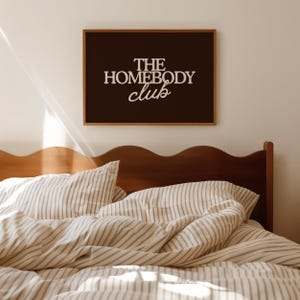 May include: A rectangular wall art print in a thin wooden frame featuring a dark brown background with the white text THE HOMEBODY club centered in a mix of bold serif and script fonts, positioned above a wooden headboard and striped bedding.