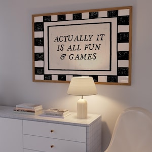 K&ouml;nnte beinhalten: Ein gerahmtes Wandkunstwerk mit dem Text "ACTUALLY IT IS ALL FUN & GAMES" h&auml;ngt &uuml;ber einem wei&szlig;en Schrank. Das Kunstwerk hat einen schwarz-wei&szlig; karierten Rand. Eine wei&szlig;e Lampe und B&uuml;cher stehen auf dem Schrank. Ein wei&szlig;er Stuhl befindet sich im Vordergrund.