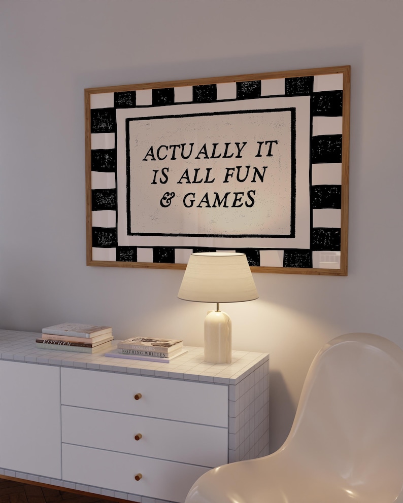 K&ouml;nnte beinhalten: Ein gerahmtes Wandkunstwerk mit dem Text "ACTUALLY IT IS ALL FUN & GAMES" h&auml;ngt &uuml;ber einem wei&szlig;en Schrank. Das Kunstwerk hat einen schwarz-wei&szlig; karierten Rand. Eine wei&szlig;e Lampe und B&uuml;cher stehen auf dem Schrank. Ein wei&szlig;er Stuhl befindet sich im Vordergrund.