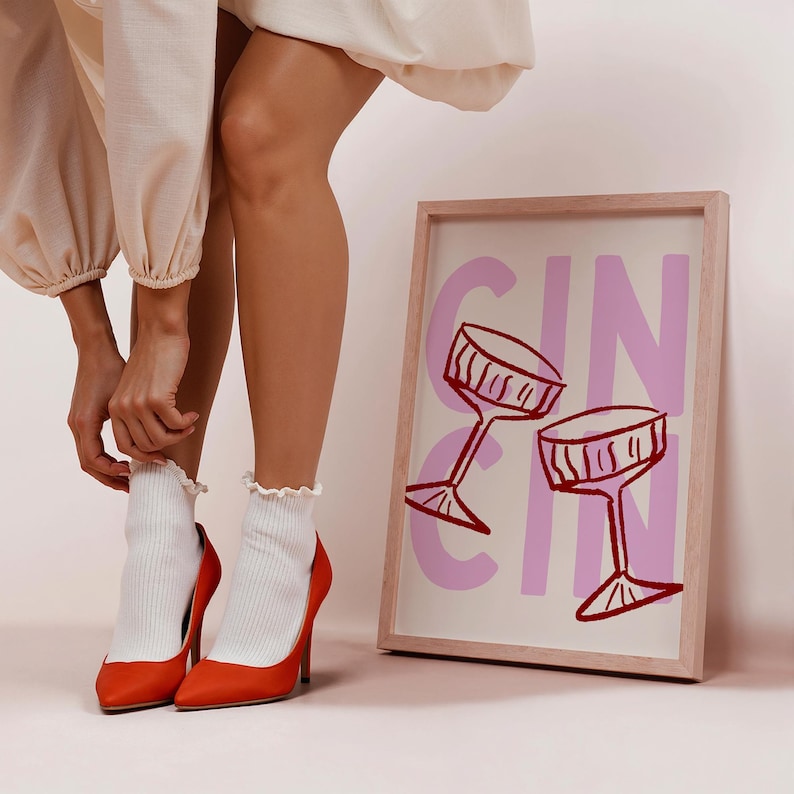Puede incluir: Un cuadro enmarcado con la palabra "GIN" en rosa y dos copas de c&oacute;ctel rojas. La imagen est&aacute; apoyada contra una pared. La persona lleva tacones altos naranjas y calcetines blancos.