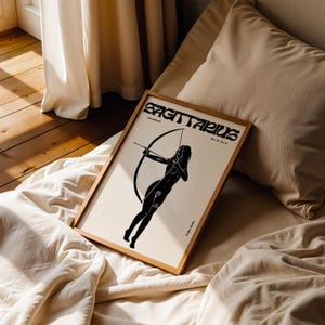 May include: A framed black and white illustration of a Sagittarius zodiac sign. The illustration depicts a woman with an arrow and bow. The frame is made of wood and has a light brown finish. The text "Sagittarius" is printed in black at the top of the illustration.
