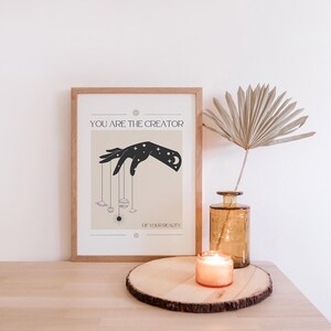 Puede incluir: Un p&oacute;ster beige con una silueta negra de una mano sosteniendo planetas y estrellas. El texto en el p&oacute;ster dice "T&uacute; eres el creador de tu realidad."