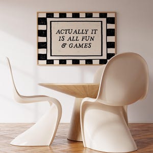 K&ouml;nnte beinhalten: Ein gerahmter Kunstdruck mit dem Text "ACTUALLY IT IS ALL FUN & GAMES" h&auml;ngt &uuml;ber einem Holztisch und zwei cremefarbenen St&uuml;hlen. Der Druck hat einen schwarz-wei&szlig;en karierten Rand.
