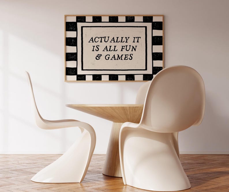 K&ouml;nnte beinhalten: Ein gerahmter Kunstdruck mit dem Text "ACTUALLY IT IS ALL FUN & GAMES" h&auml;ngt &uuml;ber einem Holztisch und zwei cremefarbenen St&uuml;hlen. Der Druck hat einen schwarz-wei&szlig;en karierten Rand.