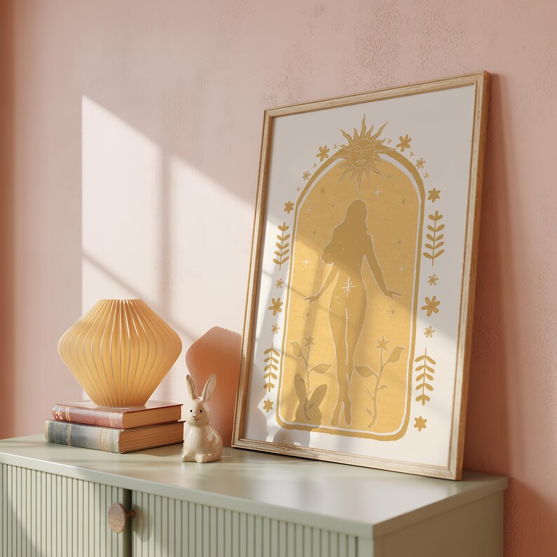 May include: A framed vertical wall art print featuring a mustard yellow archway design. Inside the arch is a silhouette of a person standing among flowers, topped with a sun icon. The print is displayed in a light wood frame on a green cabinet.