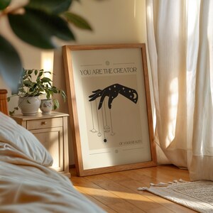 Puede incluir: Un p&oacute;ster beige con una silueta negra de una mano sosteniendo planetas y estrellas. El texto "YOU ARE THE CREATOR OF YOUR REALITY" est&aacute; impreso en el p&oacute;ster.