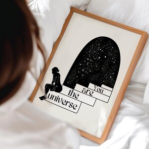 Puede incluir: Ilustraci&oacute;n en blanco y negro de una persona sentada en una luna creciente con estrellas en el fondo. El texto "You are the universe" est&aacute; escrito en la imagen.