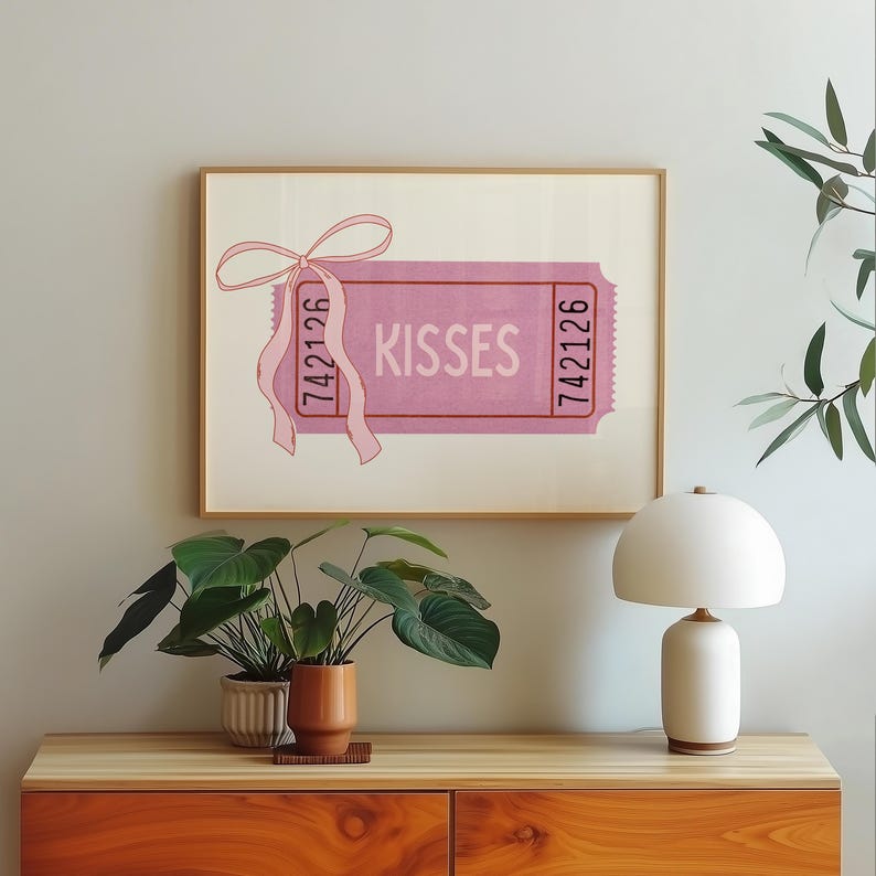May include: A framed pink 'Kisses' ticket art print. The ticket features the word 'Kisses' in white, with the numbers '742126' on each end. A pink ribbon is tied to the top left corner. The artwork is displayed on a wooden cabinet with plants and a white lamp.