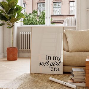 Puede incluir: Un cuadro enmarcado con el texto "In my soft girl era." El cuadro se exhibe en una habitaci&oacute;n luminosa con una ventana, una planta en maceta y un sof&aacute; beige. El marco tiene un acabado dorado.