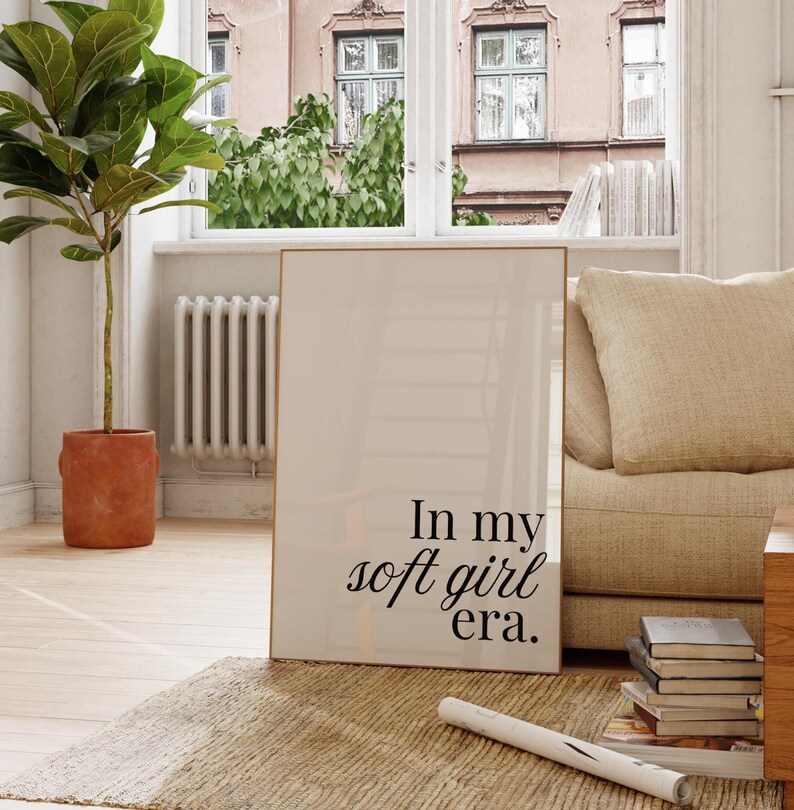 Puede incluir: Un cuadro enmarcado con el texto "In my soft girl era." El cuadro se exhibe en una habitaci&oacute;n luminosa con una ventana, una planta en maceta y un sof&aacute; beige. El marco tiene un acabado dorado.