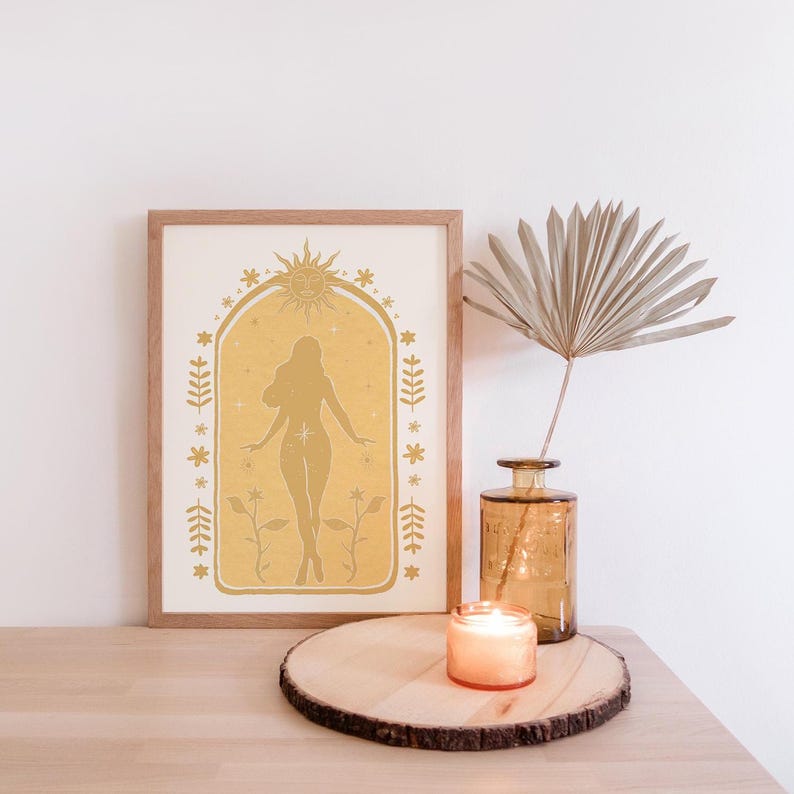 May include: A framed print featuring a mustard yellow silhouette of a person standing within an arched border, surrounded by sun, star, and floral motifs. Beside it sits a clear amber glass vase with a dried palm leaf and a small lit candle.
