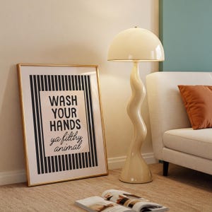 May include: A cream-coloured floor lamp with a wavy stem and a dome-shaped shade stands beside a framed print. The print has a gold frame and features the text "WASH YOUR HANDS ya filthy animal" in black and white.