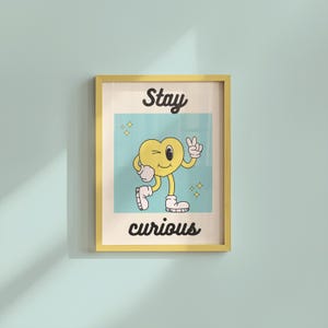 May include: A framed print with a yellow border. The artwork features a cartoon heart character winking and giving a peace sign. The words "Stay curious" are written in black script above and below the heart.