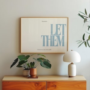 May include: A framed print with the words "LET THEM" in light blue, with the text "Reminder" and "Focus on your own path". The print hangs above a wooden cabinet with plants and a white lamp.