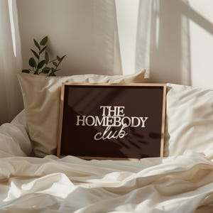 May include: A rectangular framed wall art piece featuring a dark brown background with the white text THE HOMEBODY club centered in the middle. The frame is made of light-colored wood and is positioned against a soft, draped white fabric surface.