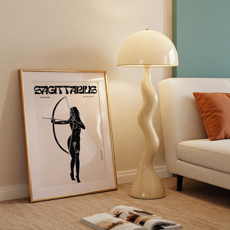 May include: A gold framed poster with a black and white illustration of a woman with a bow and arrow. The poster says "Sagittarius" in bold black letters. The poster is leaning against a white wall next to a cream colored lamp with a mushroom shaped shade.