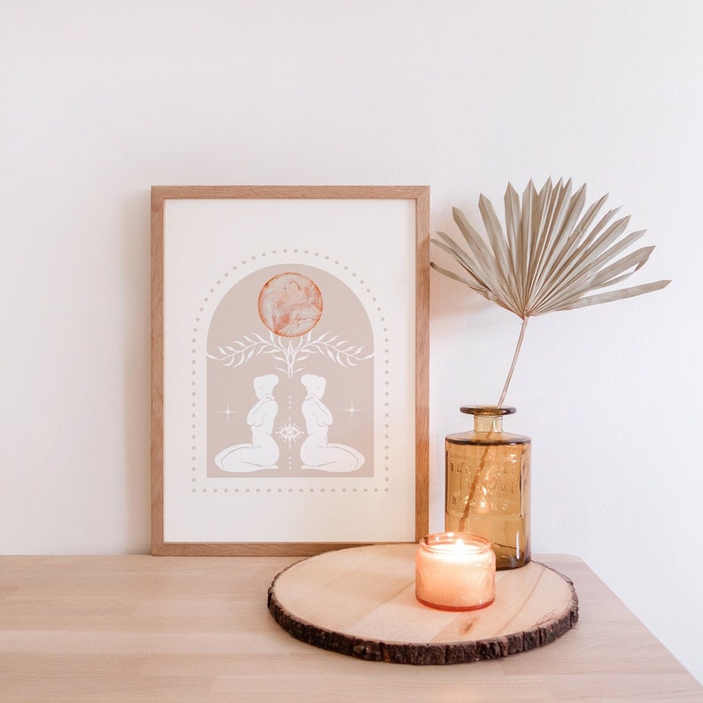 K&ouml;nnte beinhalten: Ein gerahmter Druck mit beigefarbenem Hintergrund, der zwei Figuren zeigt, die sich gegen&uuml;berstehen, mit einem gro&szlig;en orangen Kreis dar&uuml;ber. Der Druck befindet sich auf einem Holztisch mit einer brennenden Kerze und einer Vase mit einem getrockneten Palmblatt.
