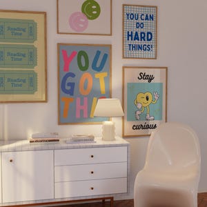 May include: A collection of framed art prints with positive messages and colorful designs. The prints include phrases like "You Got This" and "You Can Do Hard Things!" along with smiley face and cartoon character illustrations. A white cabinet and chair are also visible.
