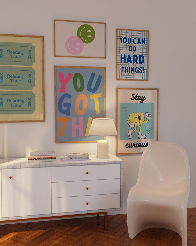 May include: A collection of framed art prints with positive messages and colorful designs. The prints include phrases like "You Got This" and "You Can Do Hard Things!" along with smiley face and cartoon character illustrations. A white cabinet and chair are also visible.