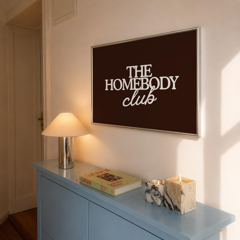 May include: A rectangular wall art piece in a silver frame featuring a dark brown background with the white text THE HOMEBODY club. The artwork is displayed above a light blue console table with a lamp, a book, and two marble candles.