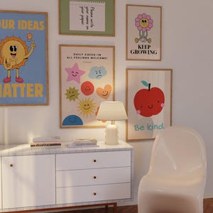 May include: A collection of framed art prints with positive messages and whimsical illustrations. The prints include a smiling sun, a flower with the text "Keep Growing", and an apple with the text "Be kind". A white cabinet and chair are in the foreground.