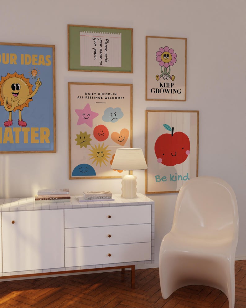May include: A collection of framed art prints with positive messages and whimsical illustrations. The prints include a smiling sun, a flower with the text "Keep Growing", and an apple with the text "Be kind". A white cabinet and chair are in the foreground.