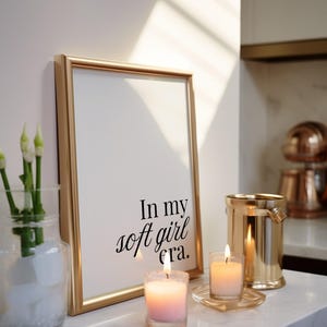 Puede incluir: Un marco dorado con el texto "In my soft girl era." est&aacute; apoyado contra una pared blanca. Dos velas encendidas y un portavelas dorado en forma de cubo est&aacute;n sobre una superficie de m&aacute;rmol blanco. Un jarr&oacute;n de cristal con flores blancas est&aacute; a la izquierda.