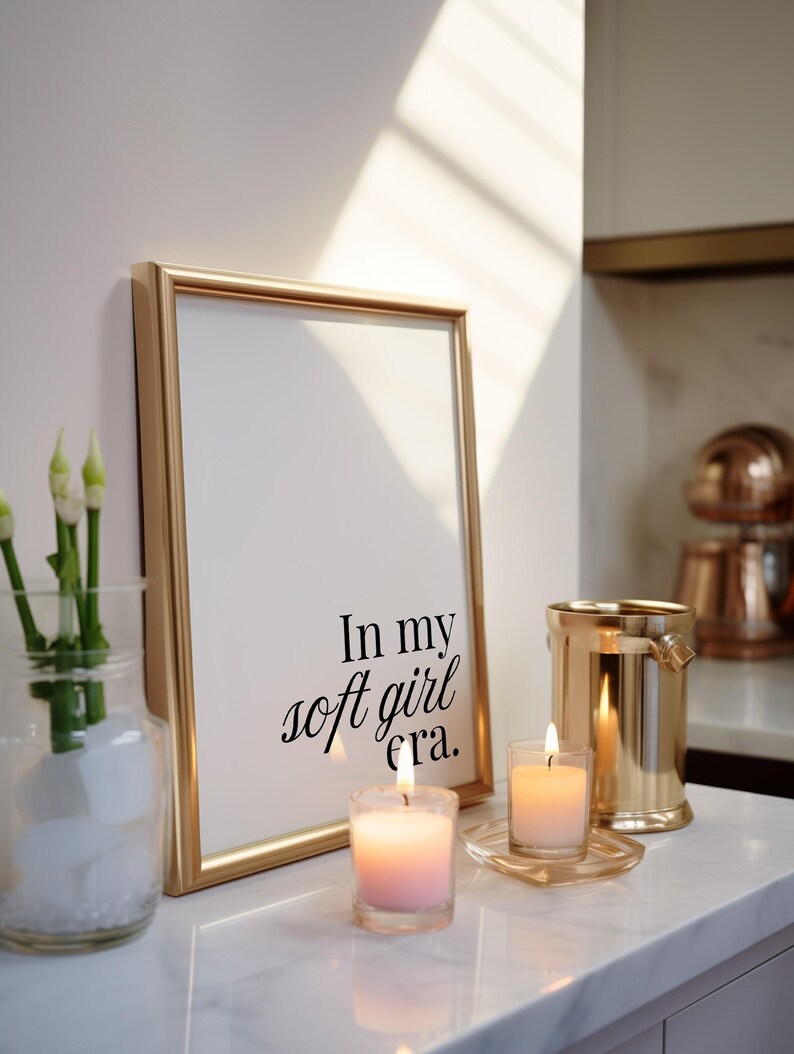 Puede incluir: Un marco dorado con el texto "In my soft girl era." est&aacute; apoyado contra una pared blanca. Dos velas encendidas y un portavelas dorado en forma de cubo est&aacute;n sobre una superficie de m&aacute;rmol blanco. Un jarr&oacute;n de cristal con flores blancas est&aacute; a la izquierda.
