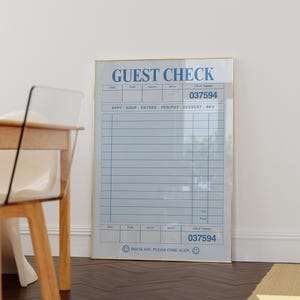 May include: A framed print featuring a light blue guest check design with the words "GUEST CHECK" at the top. The check includes sections for date, table, guests, server, and a check number. The check number is 037594. The bottom of the check reads "THANK YOU, PLEASE COME AGAIN."