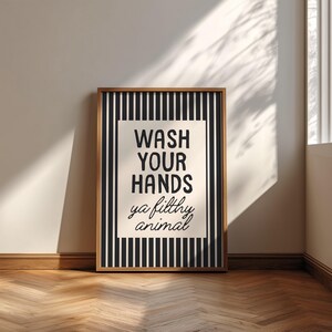 May include: A framed print with the text "WASH YOUR HANDS ya filthy animal". The artwork features a black and white striped background with a cream-colored central panel. The frame is a warm brown colour.