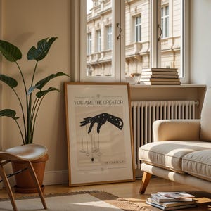 Puede incluir: Un p&oacute;ster beige con una silueta negra de una mano sosteniendo cuerdas atadas a planetas y estrellas. El texto "You are the creator of your reality" est&aacute; impreso en el p&oacute;ster.