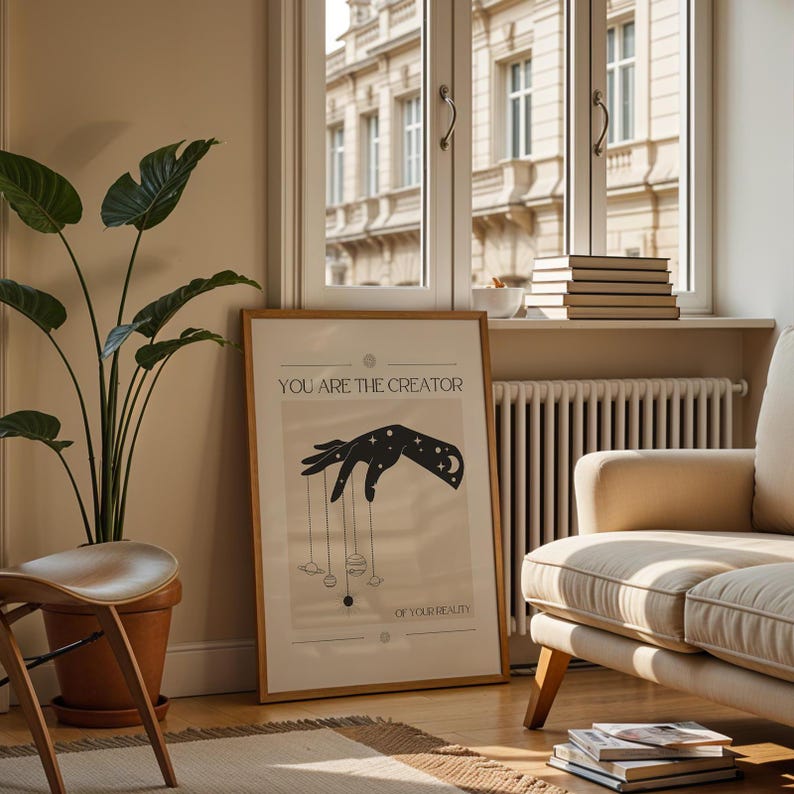 Puede incluir: Un p&oacute;ster beige con una silueta negra de una mano sosteniendo cuerdas atadas a planetas y estrellas. El texto "You are the creator of your reality" est&aacute; impreso en el p&oacute;ster.
