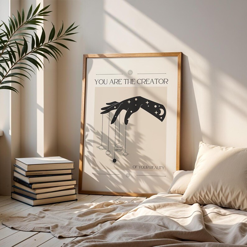 Puede incluir: Una ilustraci&oacute;n en blanco y negro de una mano sosteniendo cuerdas con planetas y estrellas unidos. El texto "You are the creator of your reality" est&aacute; debajo de la mano. La ilustraci&oacute;n est&aacute; enmarcada en un marco de madera claro.