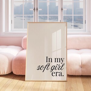 Puede incluir: Una impresi&oacute;n enmarcada con el texto "In my soft girl era." La obra de arte tiene un fondo color crema y un marco dorado. La impresi&oacute;n se exhibe en una habitaci&oacute;n con un sof&aacute; rosa y suelo de madera.