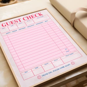 Puede incluir: Un cheque de invitado rosa en un marco de madera. Las palabras "GUEST CHECK" est&aacute;n impresas en rosa en la parte superior. El cheque incluye secciones para la fecha, la mesa, los invitados, el camarero y una lista de alimentos y bebidas. La parte inferior dice "THANK YOU. PLEASE COME AGAIN."
