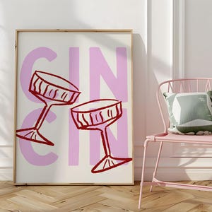 Puede incluir: Una impresi&oacute;n art&iacute;stica enmarcada con la palabra "GIN" en letras rosas grandes y dos copas de c&oacute;ctel rojas. Una silla rosa con un coj&iacute;n verde y blanco est&aacute; a la derecha de la obra de arte. La obra est&aacute; en una pared blanca.