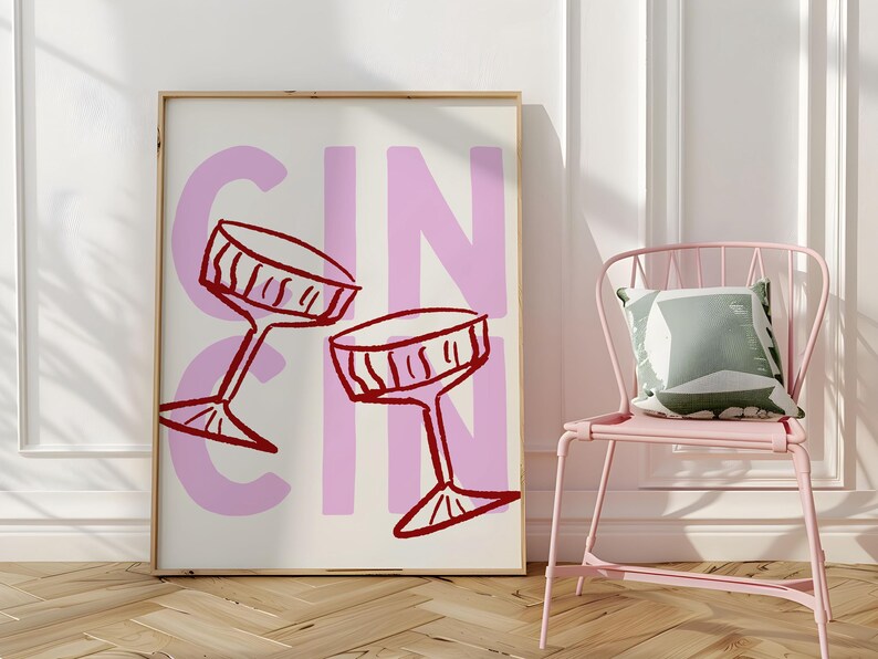 Puede incluir: Una impresi&oacute;n art&iacute;stica enmarcada con la palabra "GIN" en letras rosas grandes y dos copas de c&oacute;ctel rojas. Una silla rosa con un coj&iacute;n verde y blanco est&aacute; a la derecha de la obra de arte. La obra est&aacute; en una pared blanca.