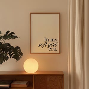 Puede incluir: Una impresi&oacute;n enmarcada con el texto "In my soft girl era" en escritura negra. La impresi&oacute;n se muestra en una pared sobre un mueble de madera, junto a una l&aacute;mpara esf&eacute;rica brillante. Tambi&eacute;n se ven una planta grande y una cortina.