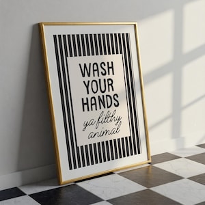 May include: A framed print with a gold border. The artwork features a black and white striped background with a central rectangular panel. The panel displays the text "WASH YOUR HANDS ya filthy animal" in black script.