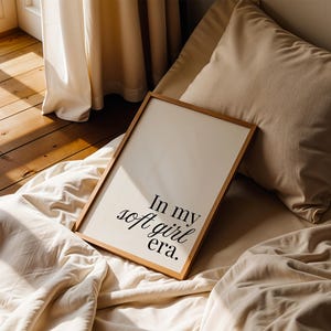 Puede incluir: Una impresi&oacute;n enmarcada con el texto "In my soft girl era" en fuente negra. La impresi&oacute;n tiene un marco de madera clara y descansa sobre una cama con ropa de cama y almohadas beige. La luz natural entra por una ventana cercana.