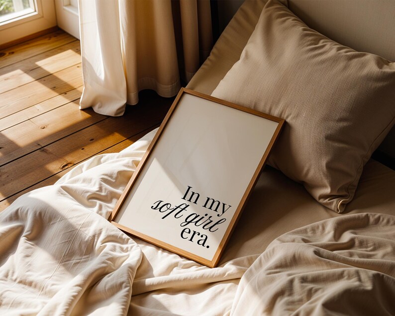 Puede incluir: Una impresi&oacute;n enmarcada con el texto "In my soft girl era" en fuente negra. La impresi&oacute;n tiene un marco de madera clara y descansa sobre una cama con ropa de cama y almohadas beige. La luz natural entra por una ventana cercana.