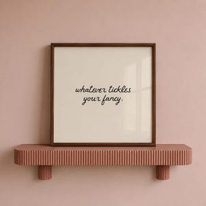May include: A square wall art print in a dark brown wooden frame featuring the black text "whatever tickles your fancy." centered on a cream-colored background. The frame is displayed on a horizontal, fluted, terracotta-colored wall shelf.