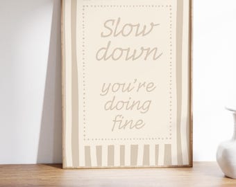 May include: A framed print with the words "Slow down you're doing fine" in a soft, neutral colour palette. The artwork features a beige background with a dotted border and vertical stripes, creating a calming aesthetic. The frame is a warm brown.