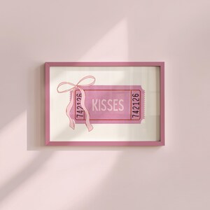 May include: A pink framed art print featuring a ticket design with the word 'KISSES' in the center. The ticket has a pink ribbon tied in a bow at the top left. The ticket has the number '742126' printed on both sides. The background is a light cream color.