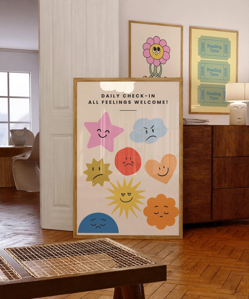 May include: A framed print with the text "DAILY CHECK-IN ALL FEELINGS WELCOME!" and colorful cartoon emotions. Additional framed prints feature a flower and "Reading Time" tickets. The room has wooden floors and furniture.
