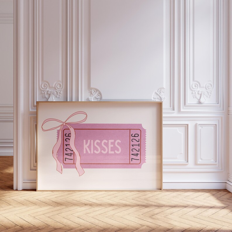 May include: A pink ticket-style art print with the word 'Kisses' in the center. The ticket has a pink bow and ribbon at the top left. The numbers '742126' are printed on both sides of the ticket. The print is framed in a light brown frame and displayed against a white wall with decorative molding.
