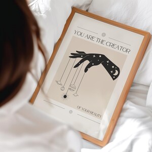 Puede incluir: Un p&oacute;ster beige con una mano negra sosteniendo cuerdas atadas a planetas. El texto en el p&oacute;ster dice "T&uacute; eres el creador de tu realidad."