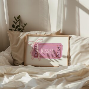 May include: A framed print featuring a pink ticket design with the word 'Kisses' in the center. The ticket has a pink ribbon attached to the top left corner. The ticket has the numbers '742126' printed on both ends. The print is displayed on a bed with white sheets and a pillow.
