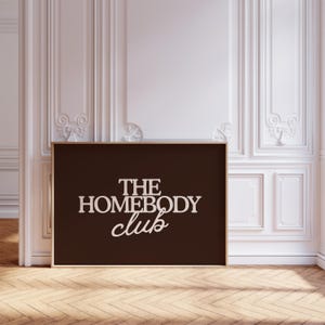 May include: A rectangular wall art print featuring a dark brown background with the text THE HOMEBODY club printed in a light beige, serif and script font combination. The print is encased in a thin, light-colored wooden frame.