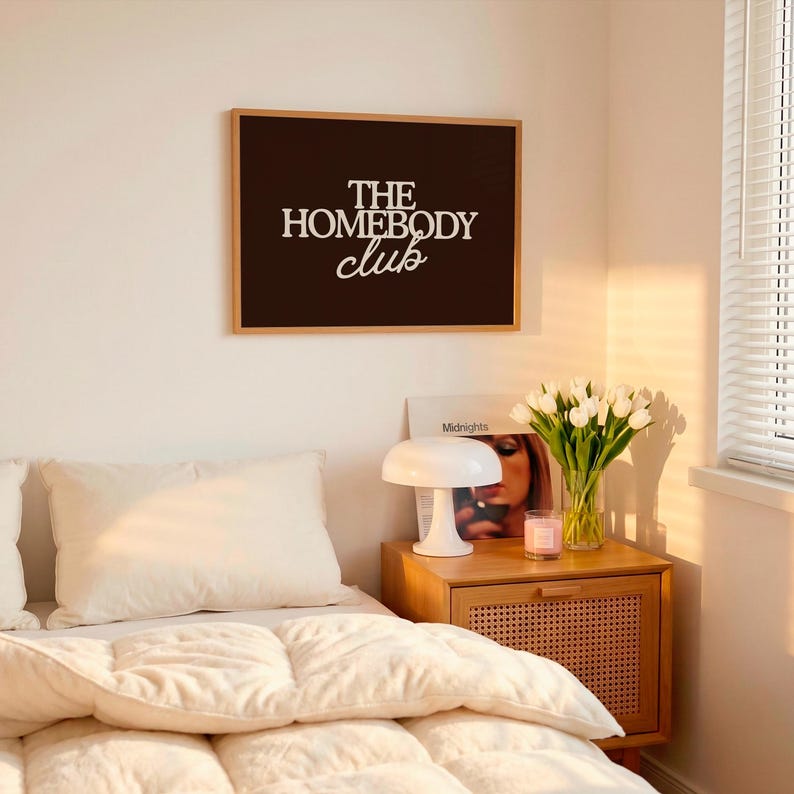 May include: A rectangular wall art print in a light wood frame featuring a dark brown background with the white text THE HOMEBODY club in a mix of bold serif and script fonts, displayed on a wall above a bedside table with a lamp and flowers.